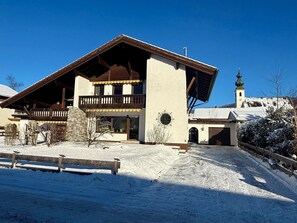 A two-story chalet with a wooden balcony, a ski lift in the background, and a snow-covered landscape.