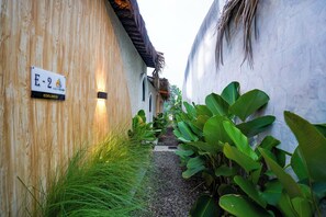 Un étroit sentier bordé de murs en bois et d’une végétation luxuriante, qui mène à un bâtiment sur lequel est apposée une plaque indiquant le numéro de la salle E-2.