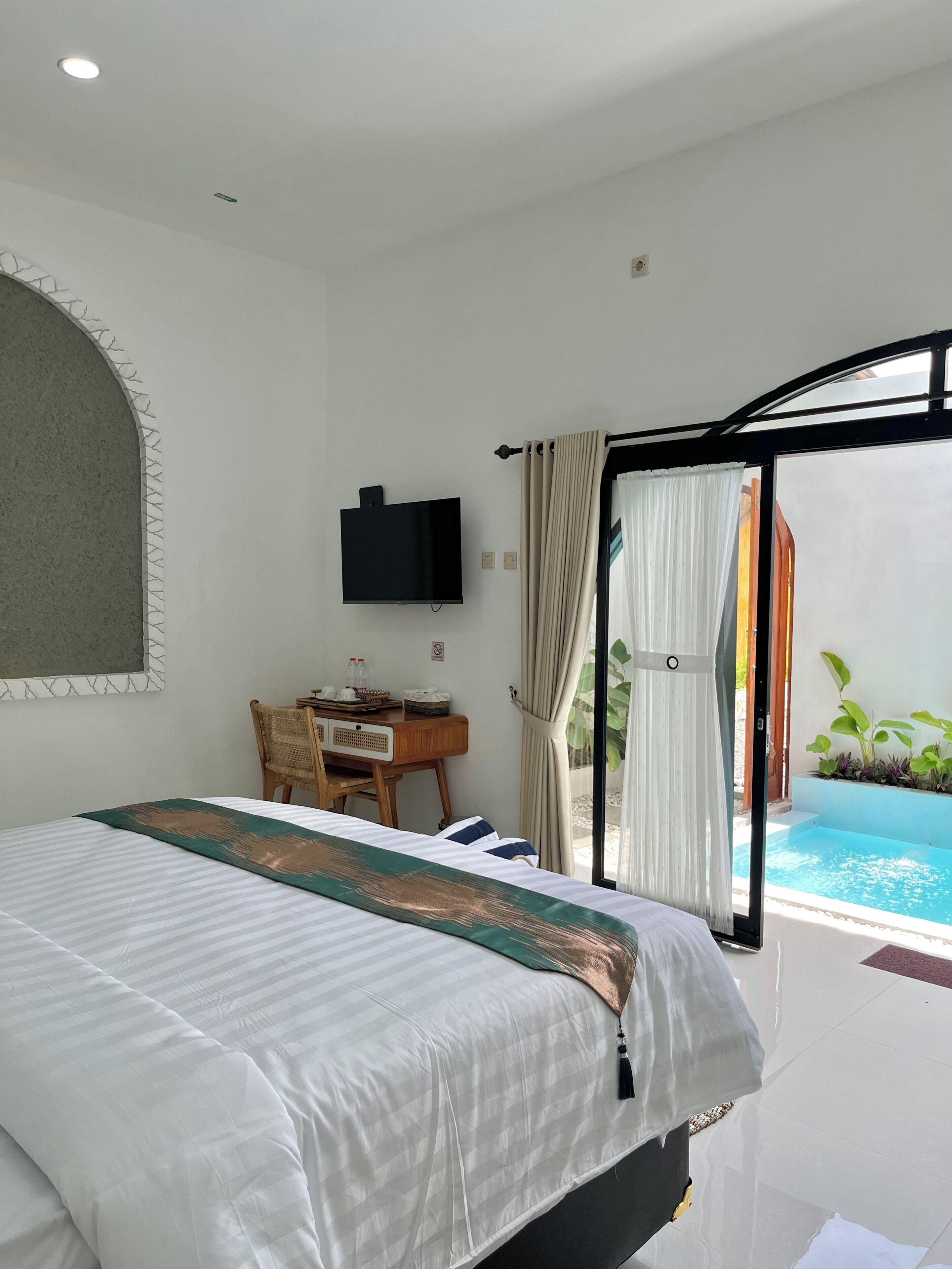 A hotel room with a bed, a desk, a TV, and a view of an outdoor swimming pool.