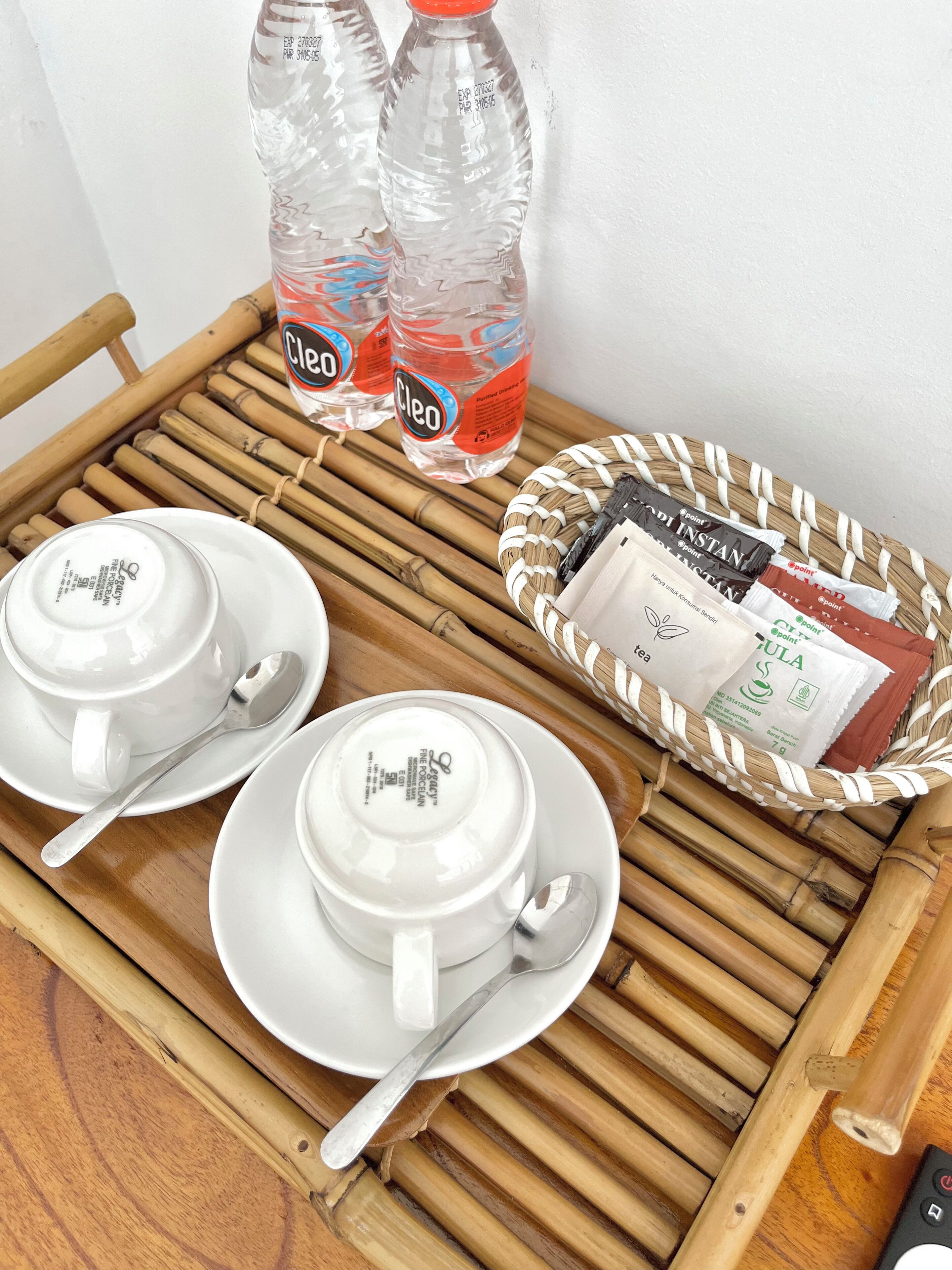 A bamboo tray with white porcelain cups, spoons, and a woven basket containing tea bags and a bottle of water.