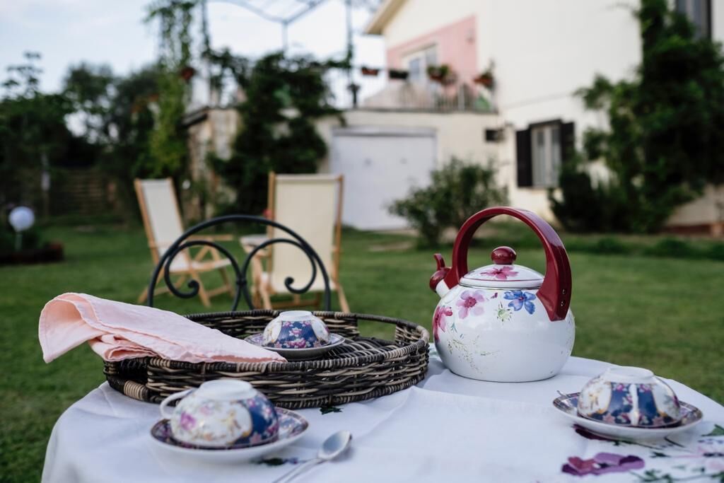 A picnic setup with a white tablecloth, a wicker basket, a floral teapot, and teacups on a grassy area.