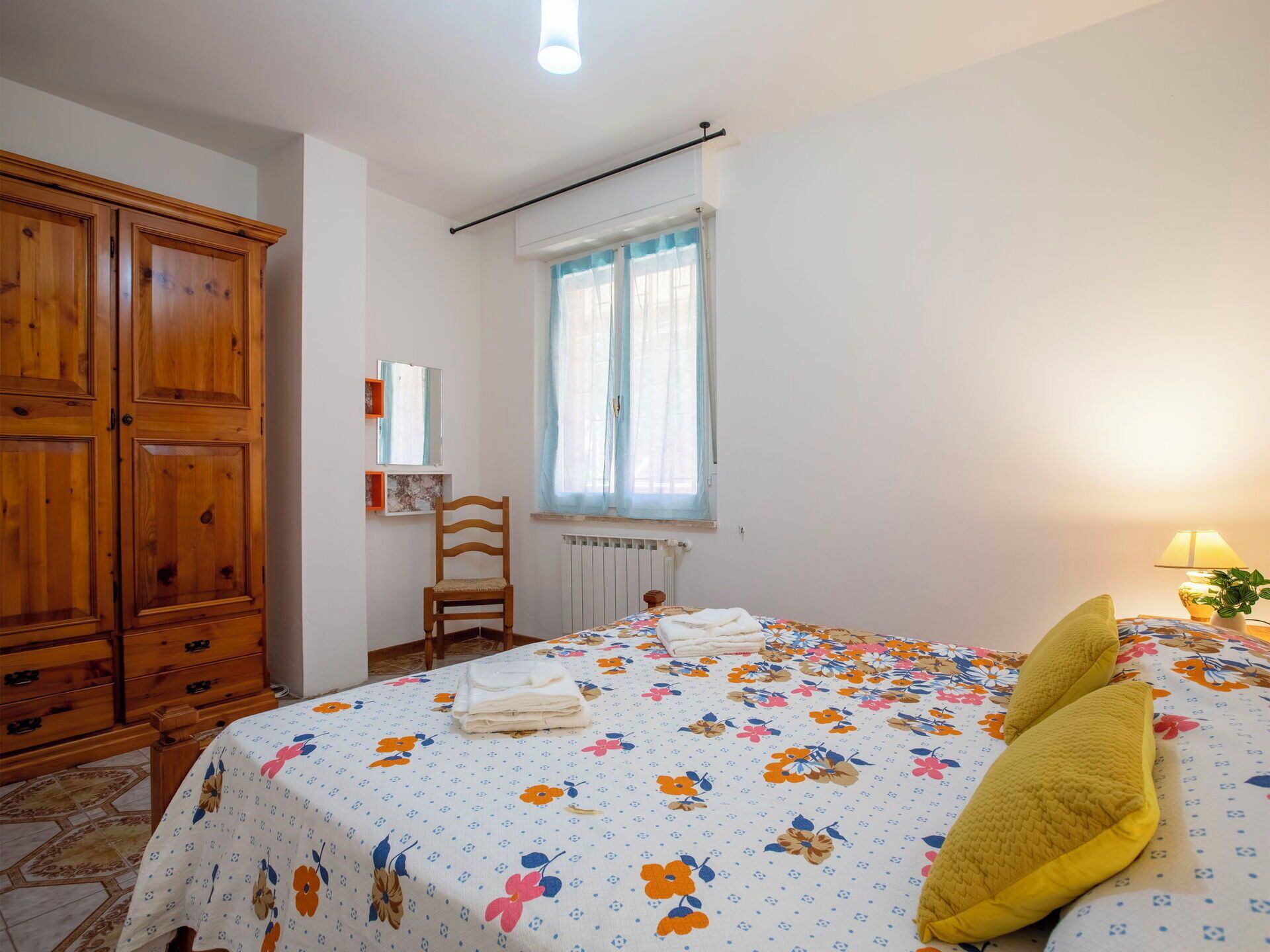 A bedroom with a bed, wooden wardrobe, chair, window, and a lamp.