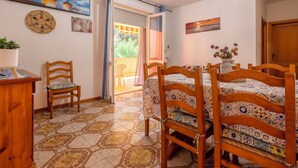 Uma área de refeições com mesa, cadeiras, uma planta em vaso e vista para um espaço exterior através de uma porta de correr.