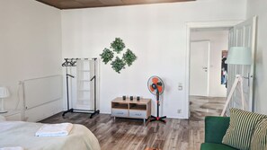 Ein Hotelzimmer mit einem Bett, einem Nachttisch aus Holz, einem Schreibtisch, einem Stuhl, einem Ventilator, einem Kleiderhaken und einer Wandpflanze.
