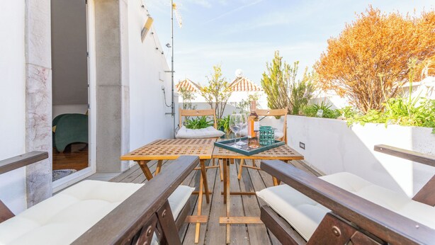 An outdoor patio with wooden furniture and a table, a door leading to a room, and a view of buildings and trees in the background. - Rosemary House (Tavira)