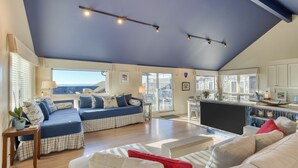 Un amplio espacio habitable con un techo alto, un ventanal con vistas, un conjunto de sofás, un televisor y una zona de cocina.
