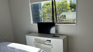 Een slaapkamer met een bed, een televisie op een wit kastje, en een raam met uitzicht op groen buiten.