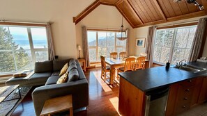 A cozy interior with a sofa, dining table, and kitchen area, featuring large windows and wooden beams.