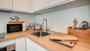 A compact kitchen with a microwave, toaster, kettle, and sink. There is a wooden cutting board with a loaf of bread and a knife. Utensils are in a wooden container. - Apartment Mika - Alpes Agency - Central Chamonix - Sleeps 4-6 (Chamonix-Mont-Blanc)