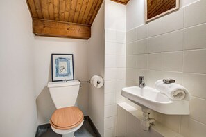 Une salle de bain avec un plafond en bois, des carreaux blancs, des toilettes avec un abattant en bois, un tableau encadré au mur et un lavabo avec une serviette.