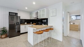 A modern kitchen with white cabinetry, a stainless steel refrigerator, a black countertop, and wooden bar stools. - Spacious 1-Bed Apartment in Coolangatta (Coolangatta)