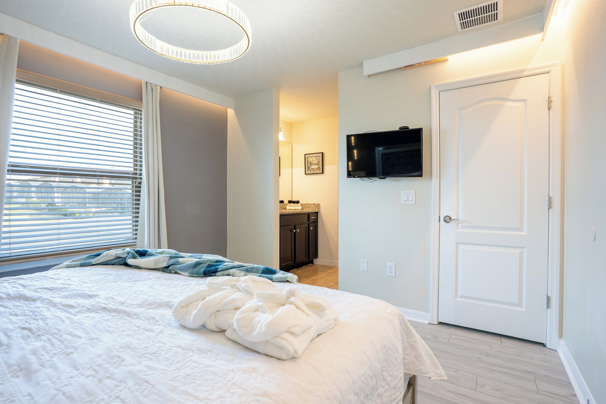 Ein Schlafzimmer mit einem Bett, einem an der Wand montierten Fernseher, einer Tür und einem Fenster mit Jalousien.