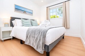 A neatly made bed with a grey blanket, a bedside table with a lamp, and a window with curtains. - Spacious 4BR Home Mins to Santa Clara & Tech Campuses (San Jose)