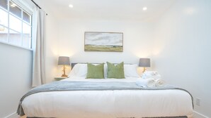 Ein ordentlich bezogenes Bett mit weißen und grauen Bettwäsche, zwei grüne Kissen und eine graue Tagesdecke. Zwei Nachttischlampen und ein Gemälde befinden sich an der Wand über dem Bett.