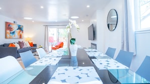 Ein moderner Essbereich mit einem Glastisch und Stühlen, einer Vase mit Blumen und einem Sofa mit orangefarbenen Kissen.