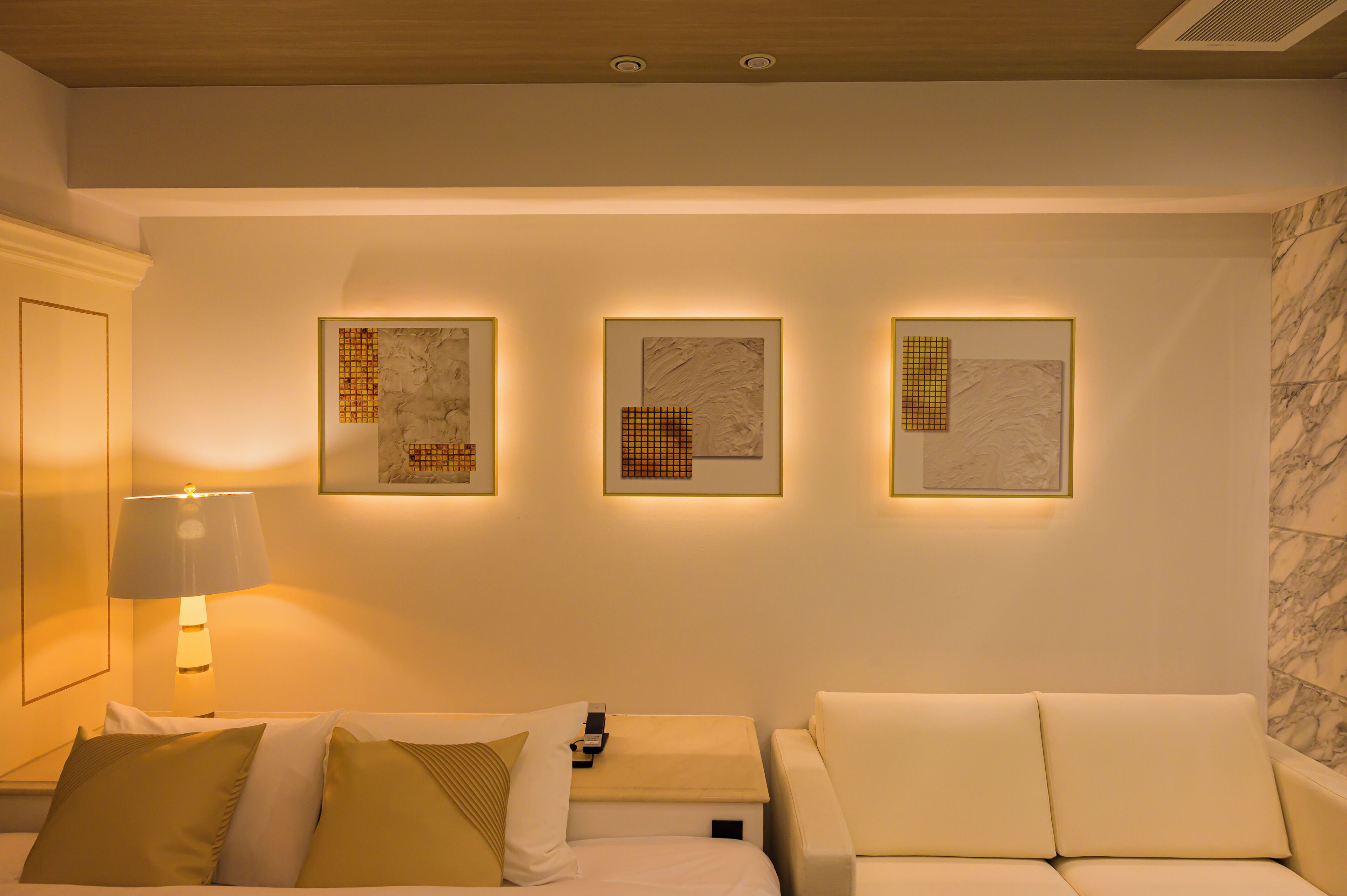A hotel room with a bed, two white sofas, a nightstand with a lamp, and three framed artworks on the wall.