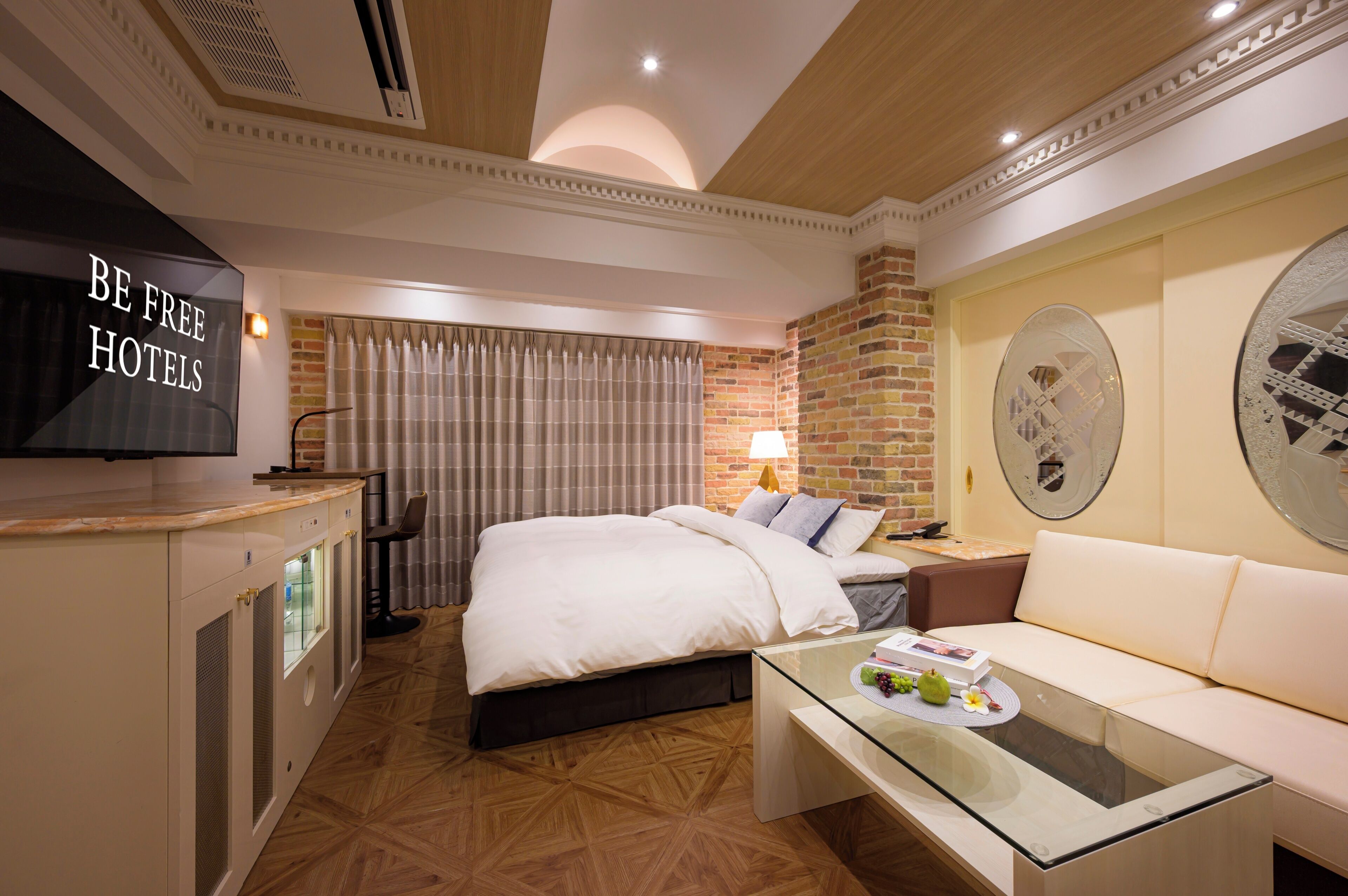 A hotel room with a bed, a sofa, a glass coffee table, a TV, and a wall with a decorative mirror.
