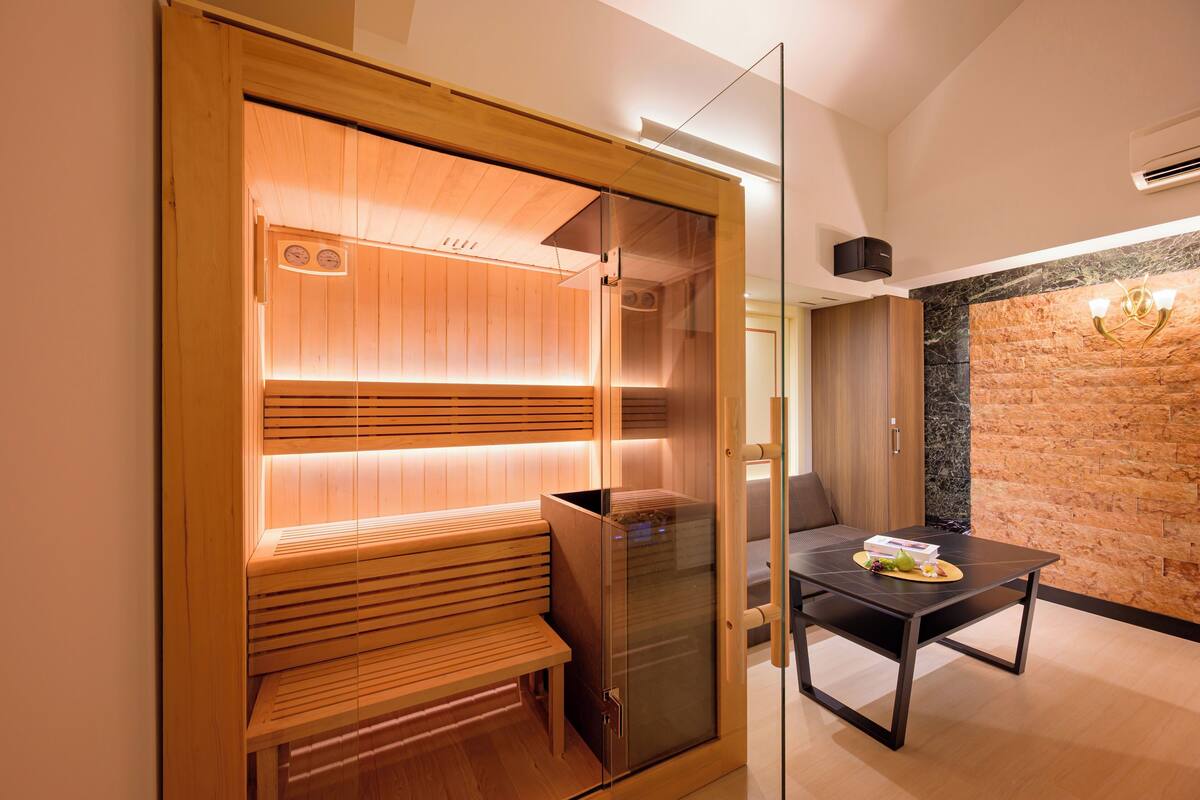 A sauna with wooden benches and a glass door, a table with a plate of fruit, and a stone wall in the background.