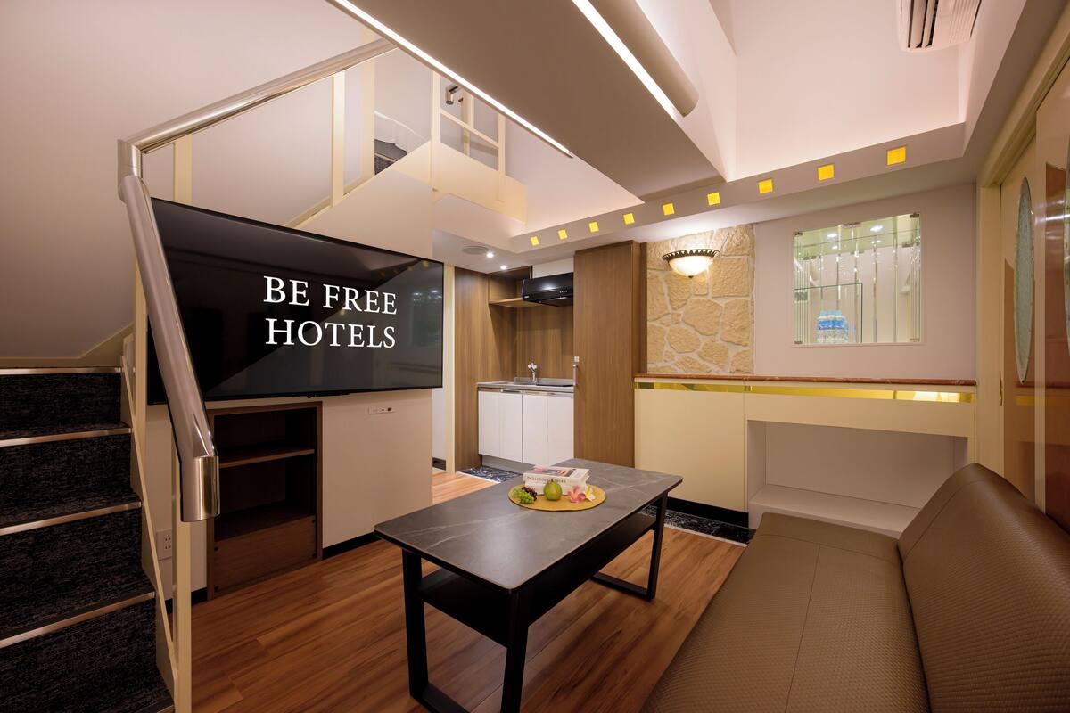 A modern hotel room with a staircase, a flat-screen TV, a dining table with fruit, a sofa, and a kitchen area.