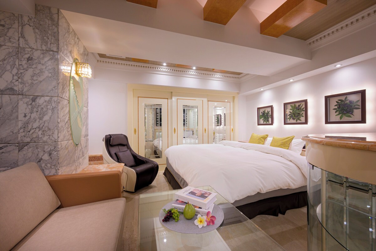 A hotel room with a marble wall, a bed with white linens, a glass coffee table with fruits and a book, a leather armchair, and a glass cabinet.