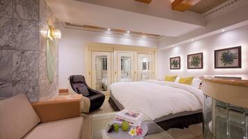 A hotel room with a marble wall, a bed with white linens, a glass coffee table with fruits and a book, a leather armchair, and a glass cabinet.
