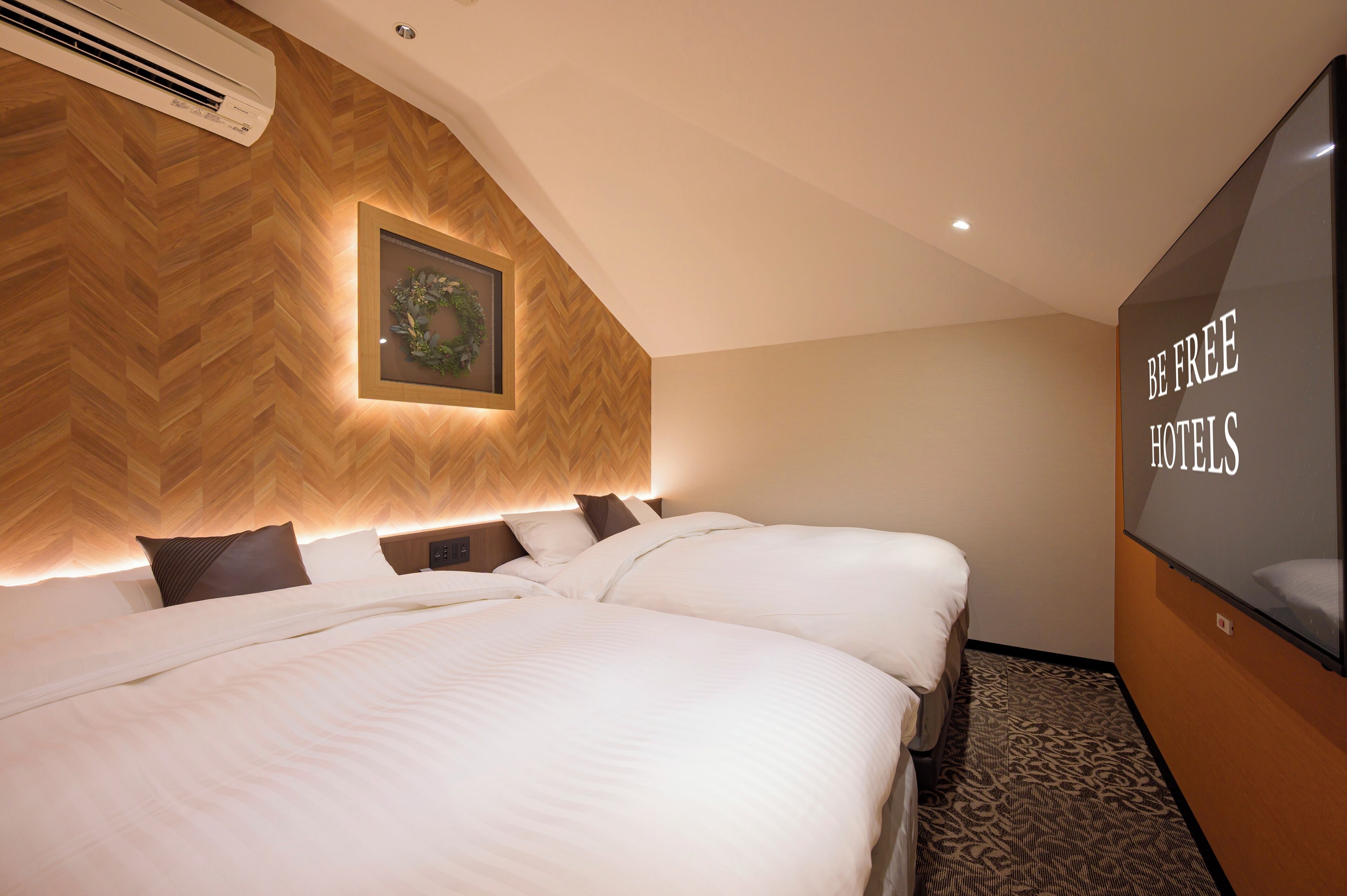 A hotel room with two beds, a wooden herringbone wall, an air conditioner, a framed picture, and a wall-mounted sign reading "BE FREE HOTELS".