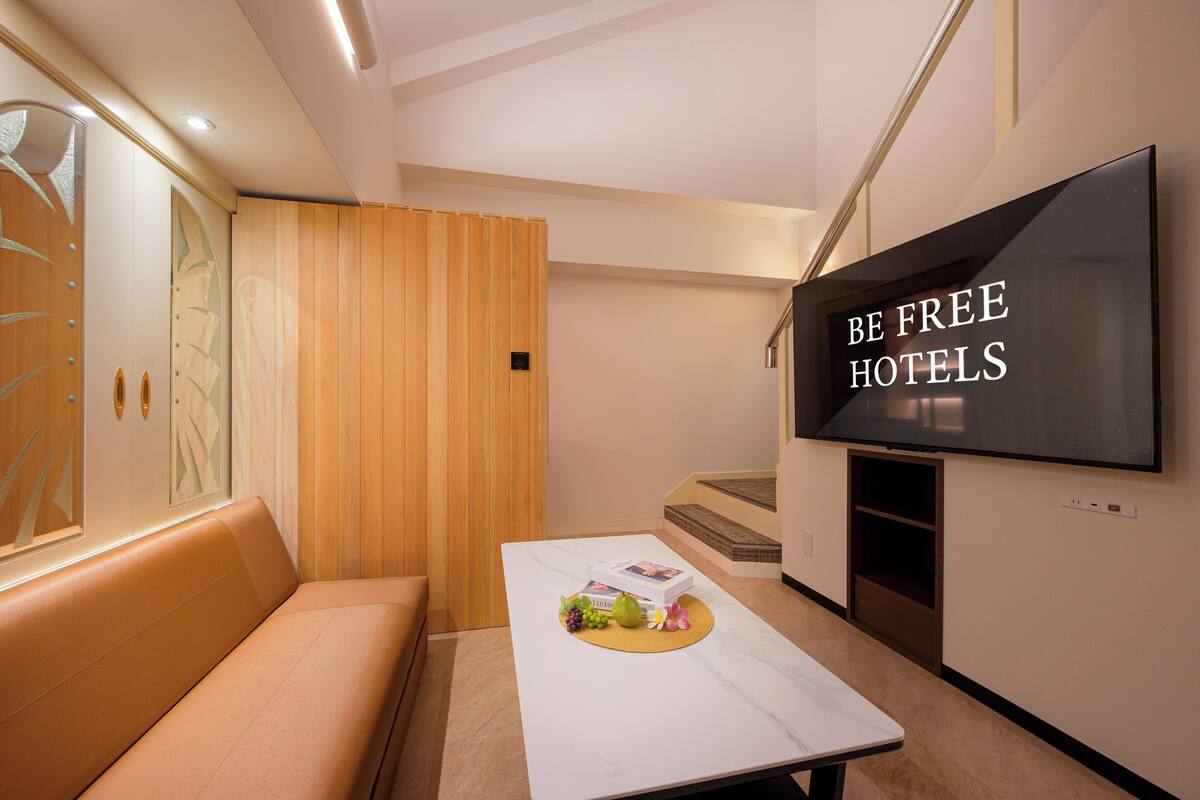 A modern hotel room with a flat-screen TV, a sofa, a coffee table with fruits and a book, and a wooden panel wall.