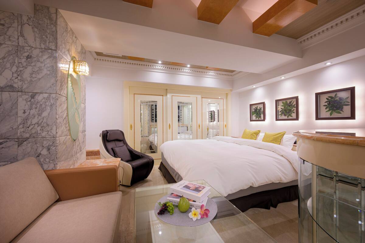 A hotel room with a marble wall, a bed with white linens, a glass coffee table with fruits and a book, a leather armchair, and a glass cabinet.