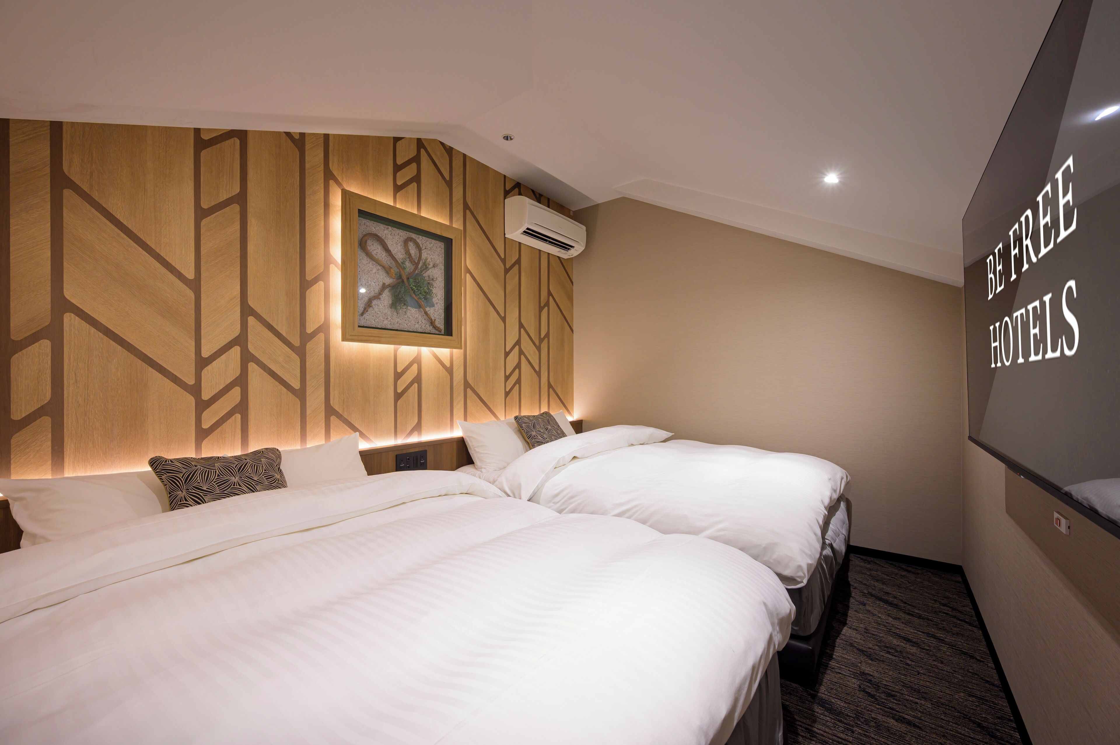 Une chambre d’hôtel avec deux lits, un climatiseur fixé au mur et une pancarte indiquant « BE FREE HOTELS ».