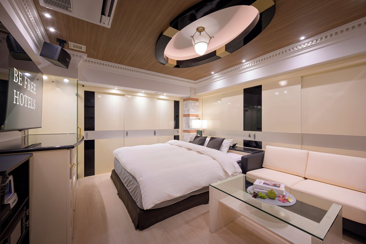 A modern hotel room with a large bed, a sofa, a glass coffee table, and a TV on a desk.
