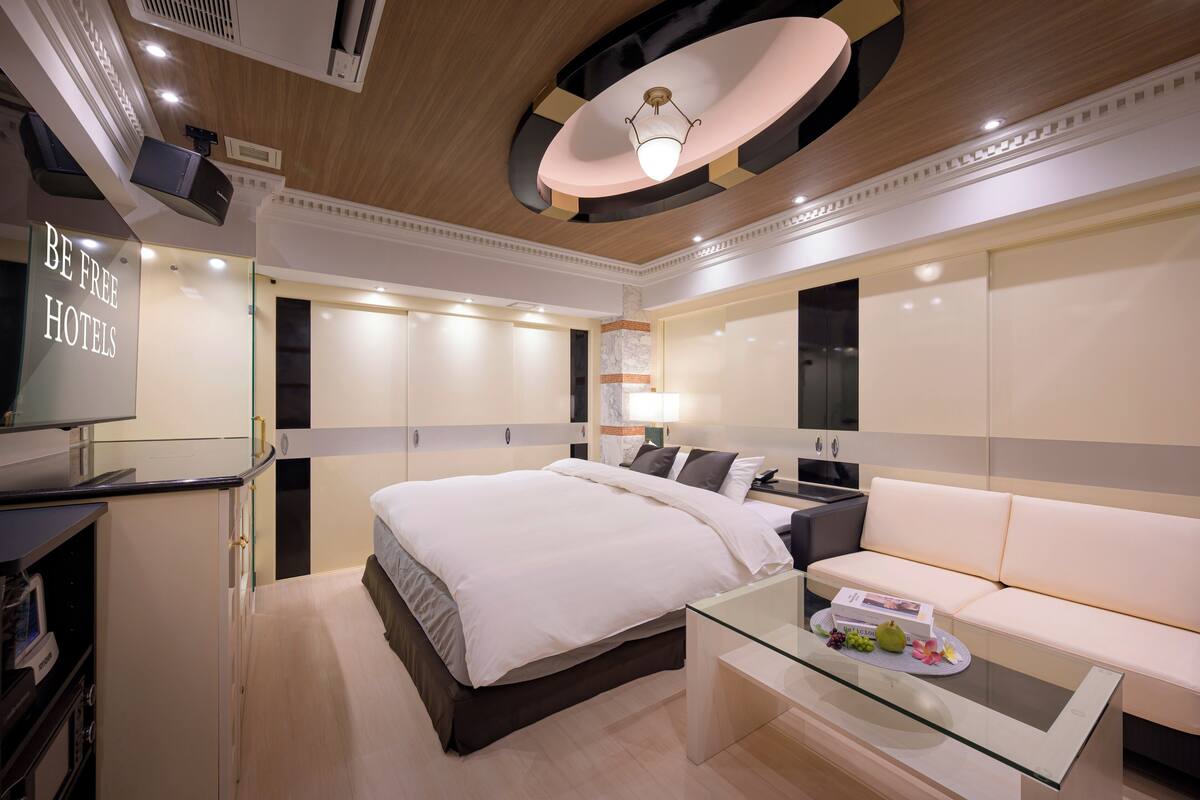 A modern hotel room with a large bed, a sofa, a glass coffee table, and a TV on a desk.