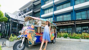 Un tuk-tuk aux couleurs vives et au design original est garé devant un bâtiment moderne doté de grandes fenêtres et de balcons.