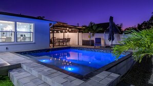 Ein moderner Poolbereich im Freien mit Terrasse und Sitzgelegenheiten, umgeben von einem Holzzaun und einem Haus mit großen Fenstern.