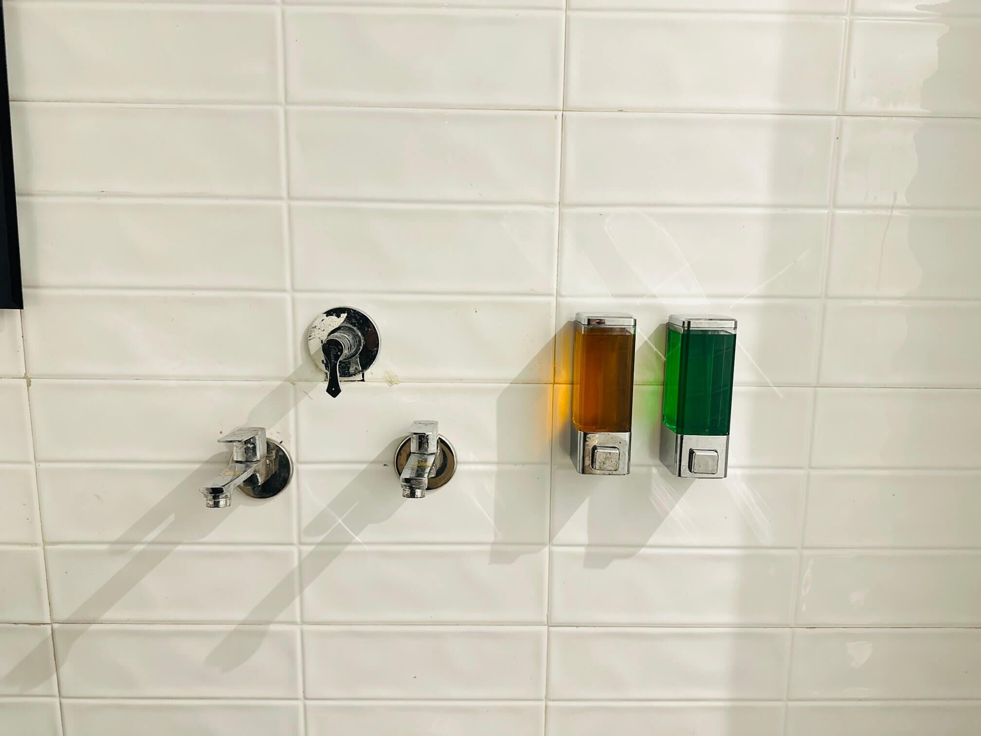 A bathroom with white tiled walls, three faucets, and two soap dispensers.