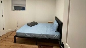 Ein kleines Zimmer mit einem Einzelbett, Holzboden, einem Fenster mit Jalousien, einer Deckenleuchte und einer Tür.