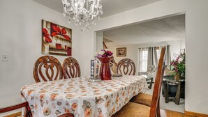 A dining area with a table covered in a floral tablecloth, surrounded by wooden chairs with heart-shaped backs. A chandelier hangs from the ceiling, and a vase with flowers is on the table. - 12 Mi to Dtwn St Paul: Extended Stay in Woodbury! (Woodbury)