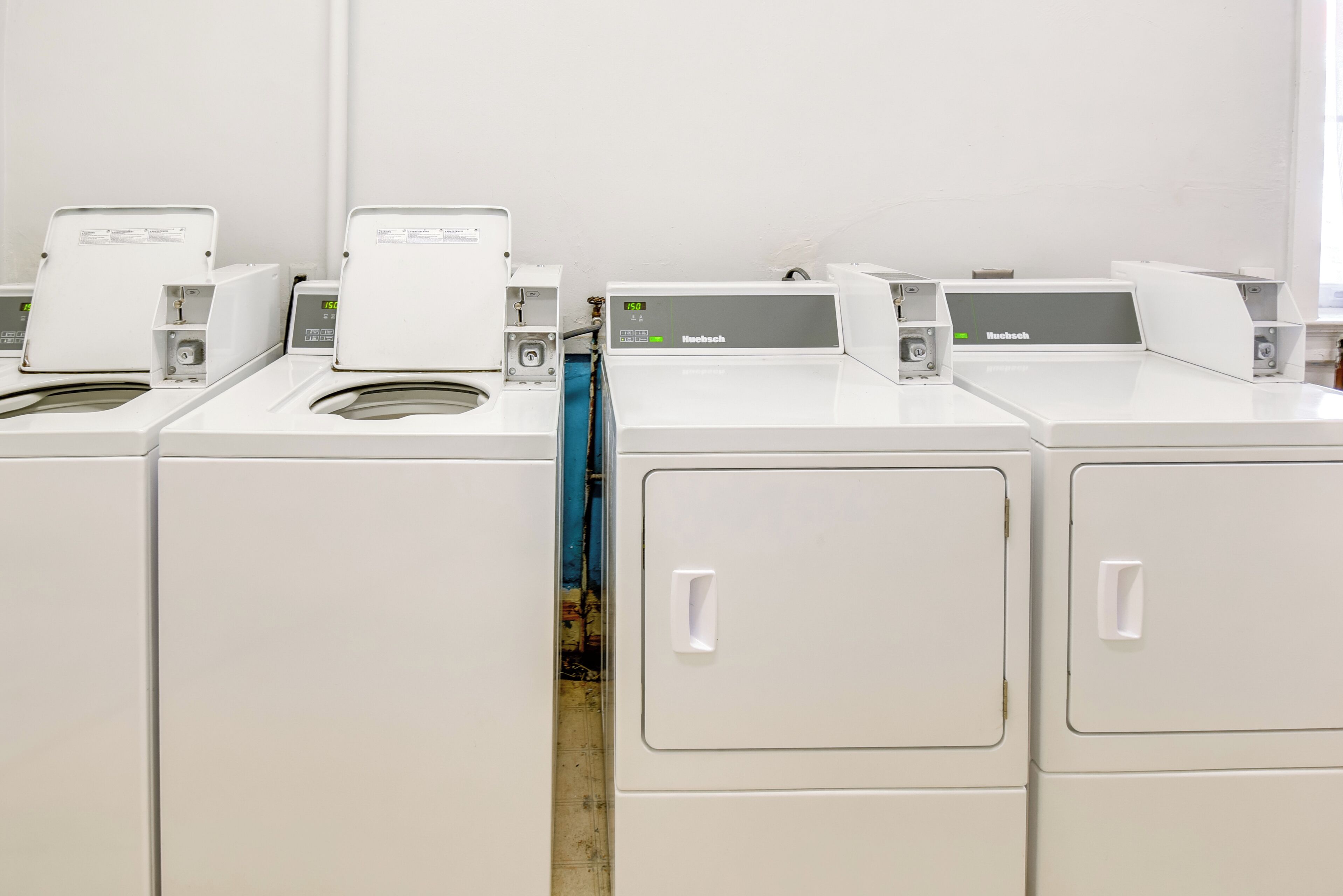 A row of white, industrial-style laundry machines with control panels, arranged in a row against a plain wall.