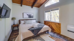 Un dormitorio con techo de madera, un ventanal grande, una cama con manta y almohadas, una mesita de noche con lámpara y un televisor montado.
