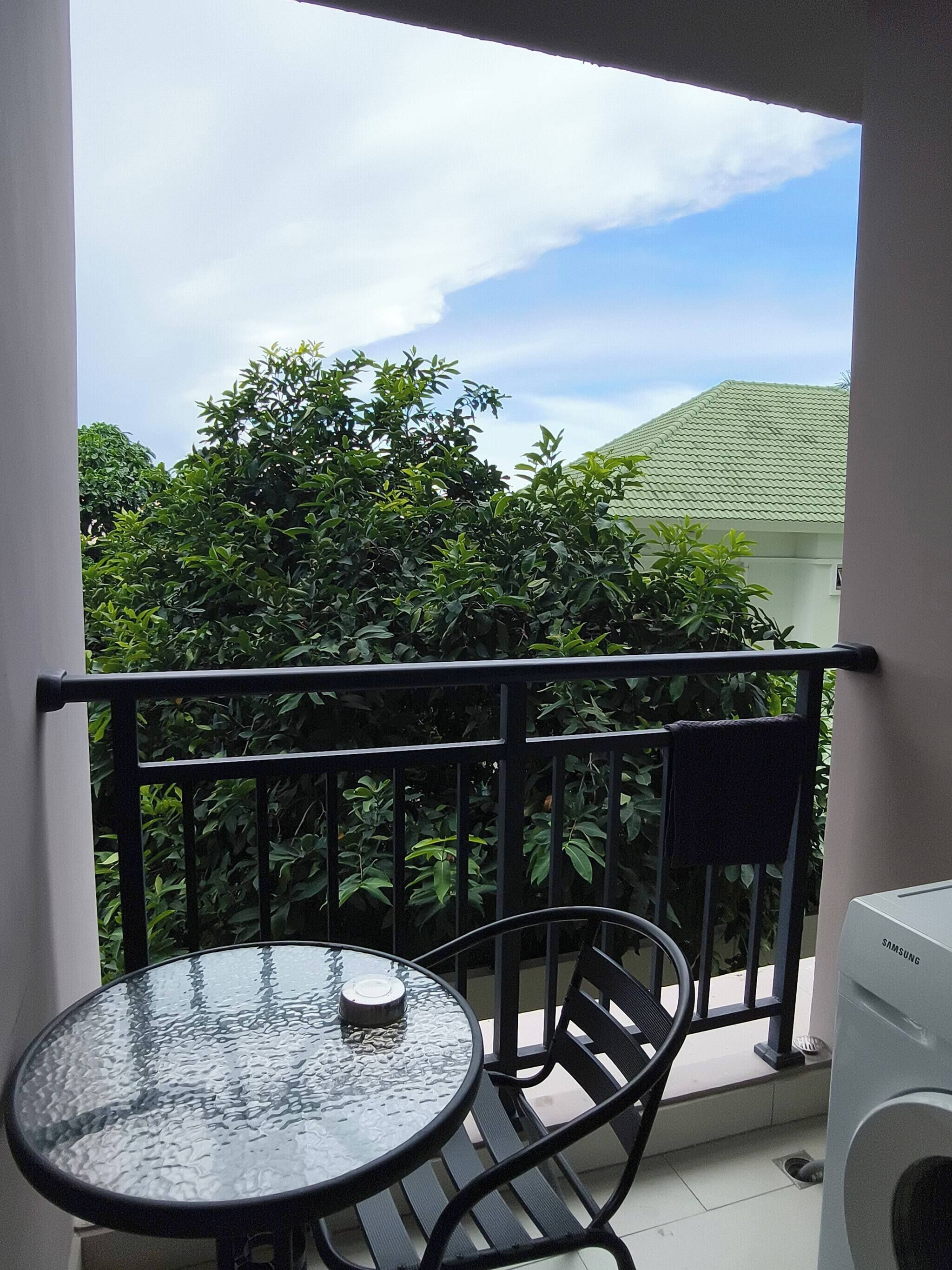 A balcony with a glass table and chairs, a washing machine, and lush greenery in the background.