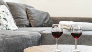 Deux verres à vin sont posés sur une table ronde dans un salon, où se trouvent un canapé gris et un coussin blanc.