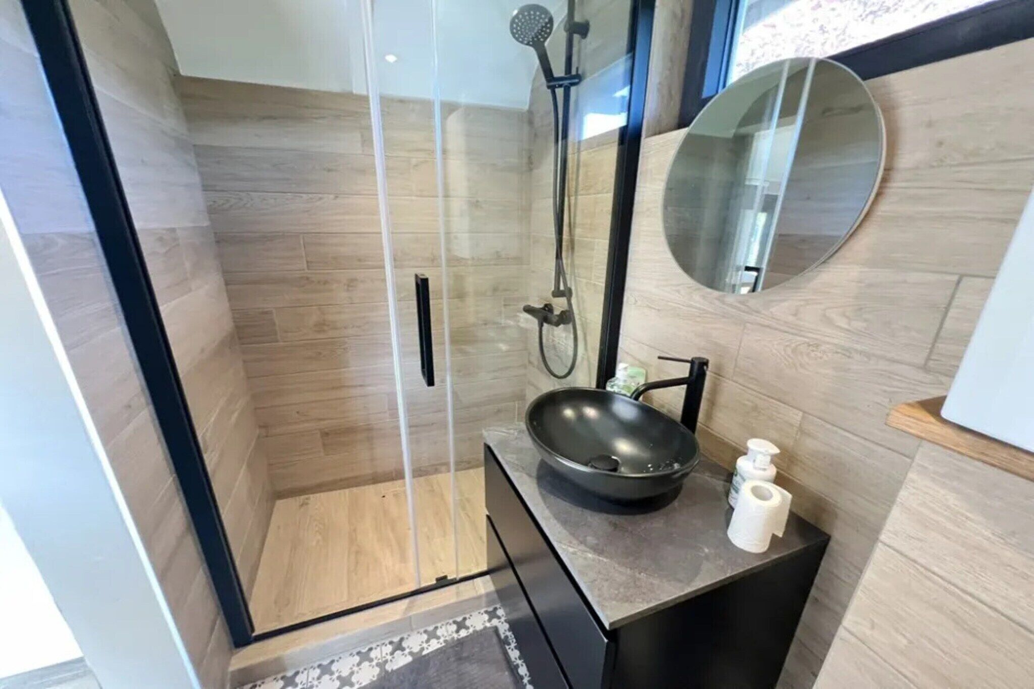 Ein modernes Badezimmer mit einer gläsernen Duschkabine, einem runden Spiegel und einem schwarzen Waschbecken auf einer dunklen Ablagefläche.