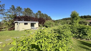 En mindre, etetages campingvogn med skråt tag, beliggende i et græsareal med tæt buskads og træer i baggrunden.