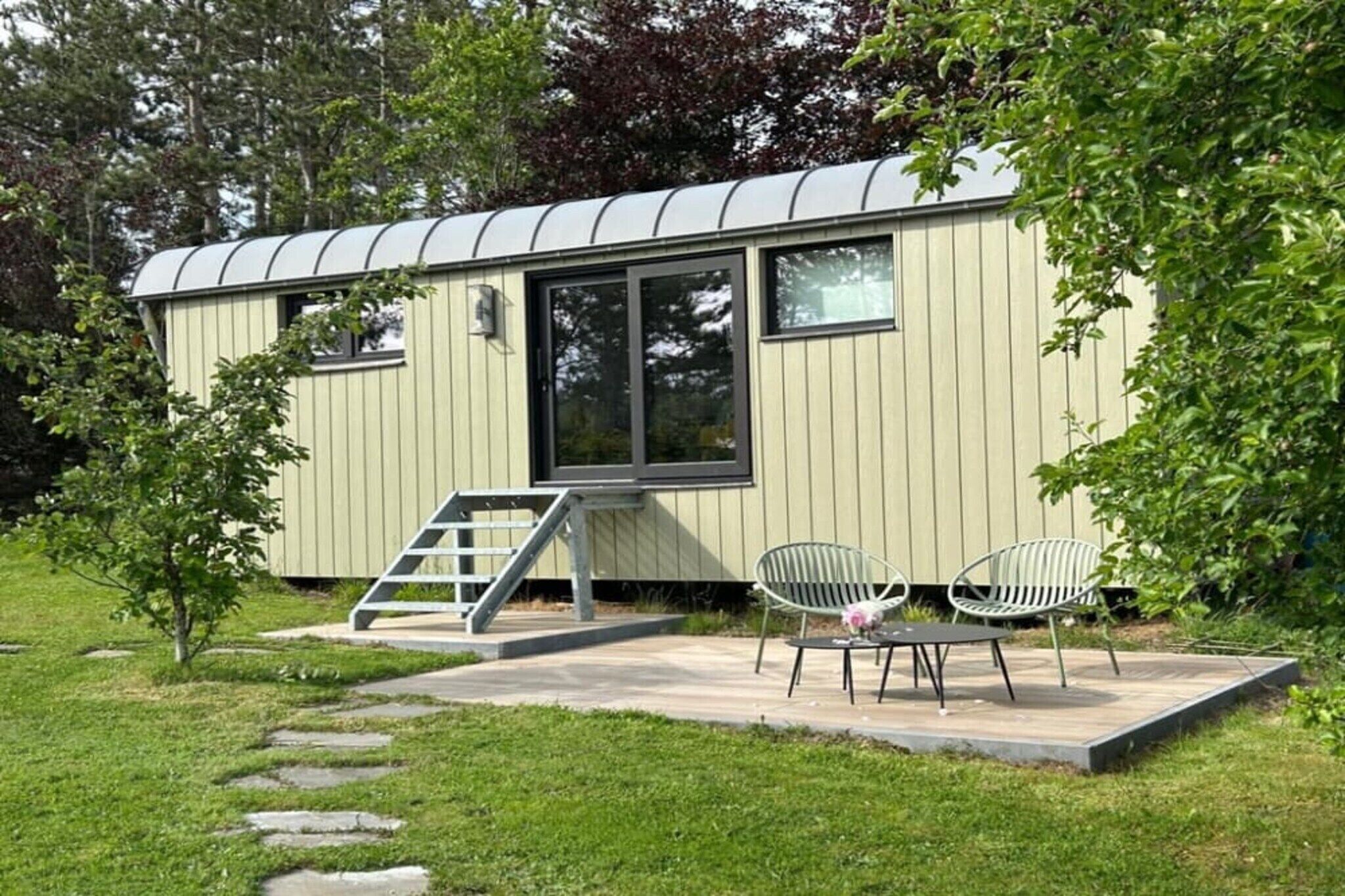 Ett enplanshus på hjul, målat i en ljusgrön färg, med ett välvt tak, ett stort fönster och en liten uteplats med två stolar och ett bord.