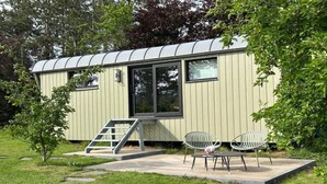 En etagers, lysegrøn campingvogn med et buet tag, et stort vindue og en lille terrasse med to stole og et bord.