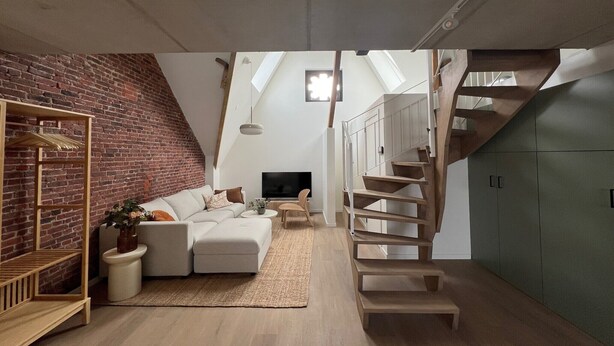 Ein Loft-Wohnzimmer mit Ziegelwand, Holztreppe, weißem Sofa und Fernseher.