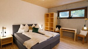 Een slaapkamer met een bed, nachtkastjes, een houten bureau en een raam met uitzicht op groen.