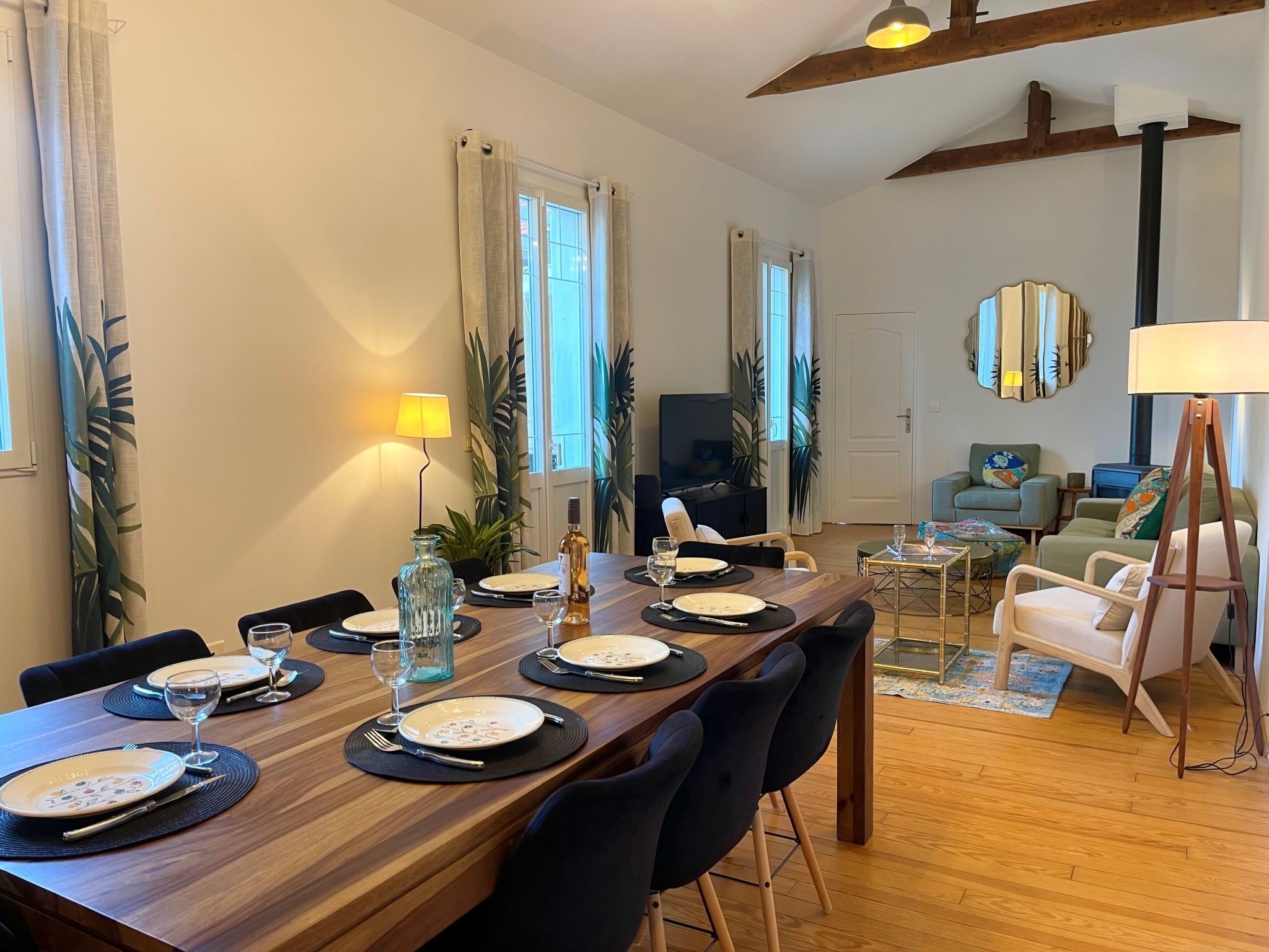 Un coin repas avec une table en bois, dressée pour quatre personnes, entourée de chaises. On aperçoit un téléviseur au fond, un canapé et une lampe.