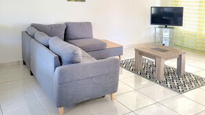 Ein modernes Wohnzimmer mit einem grauen Sofa, einem Holztisch und einem Flachbildfernseher. Drei an der Wand befestigte Kunstwerke zeigen Blätter und Pflanzen.