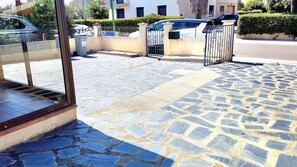 A stone-paved entrance with a metal gate, a trash bin, and a clear view of parked cars and buildings in the background. - Renovated air-conditioned house sleeps 6 5 minutes from the beach (Latour-Bas-Elne)