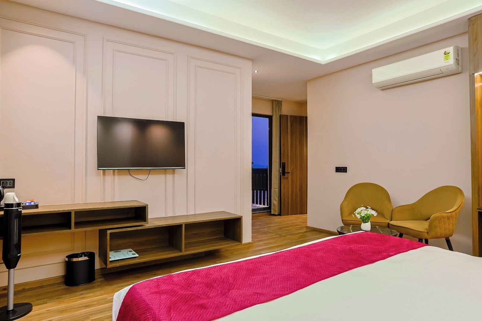 Une chambre d’hôtel avec un grand lit, un téléviseur à écran plat fixé au mur, un meuble de rangement en bois, deux fauteuils jaunes et un balcon visible à travers une porte ouverte.